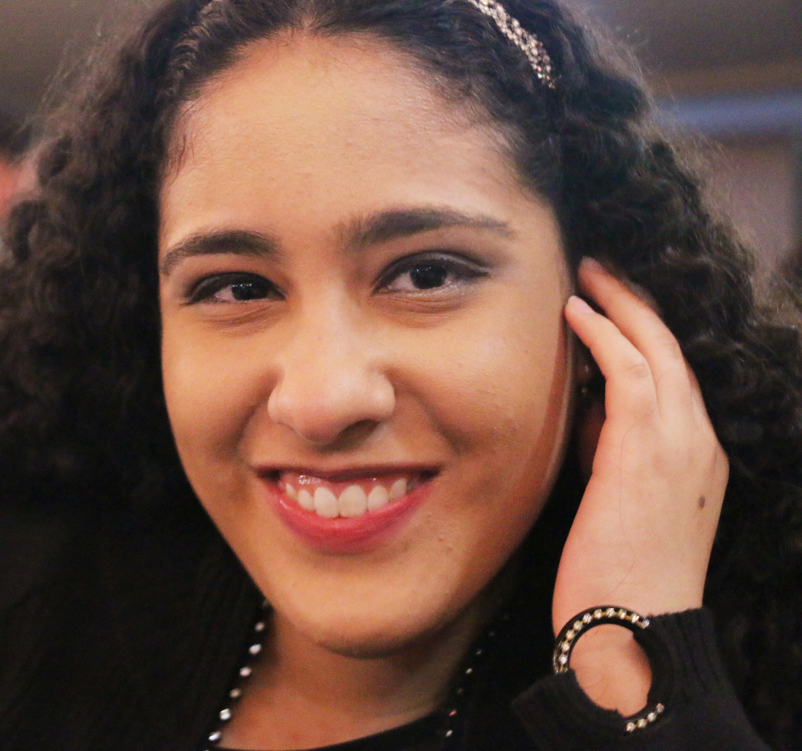 A young women with wavy black hair. She is smiling and her right hand is brushing hair away from her face and behind her ear.